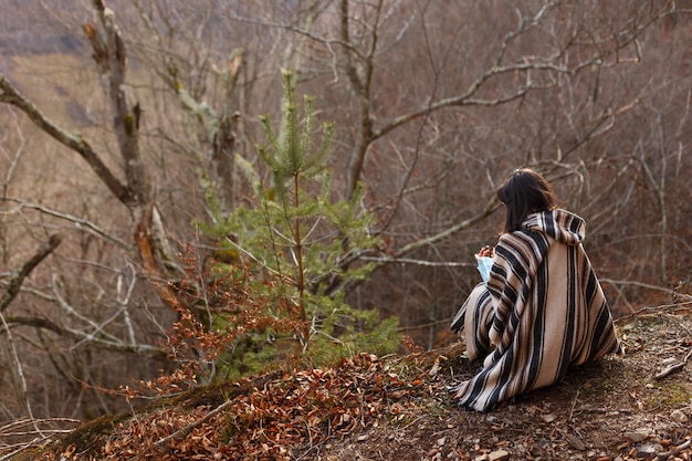 Giovane donna con capelli corti bruna in poncho seduto nei boschi autunnali, foresta.