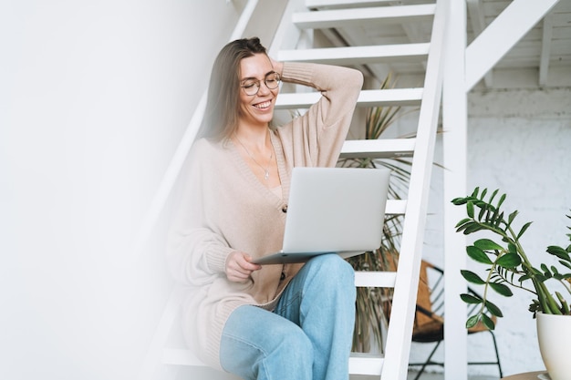 Giovane donna con capelli lunghi in cardigan che lavora al computer portatile seduto sulle scale a casa