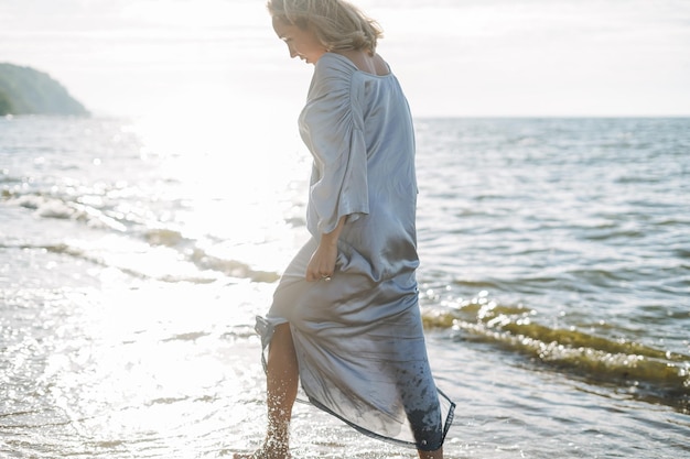 Giovane donna con capelli lunghi in vestito blu di seta che gode della vita sulla spiaggia del mare