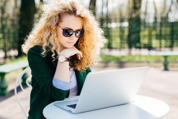 Giovane donna con capelli ricci biondi che indossa occhiali da sole ed elegante giacca verde seduta davanti a un computer portatile aperto all'aperto leggendo notizie online con espressione pensierosa Concetto di tecnologia delle persone