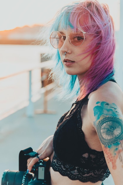 Giovane donna con capelli rosa e blu e tatuaggi che tengono una macchina fotografica in piedi vicino al mare