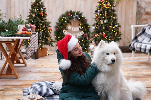 Giovane donna con cappello da Babbo Natale sullo sfondo dell'albero di Natale con cane samoiedo bianco all'aperto Decorazione del cortile per il nuovo anno