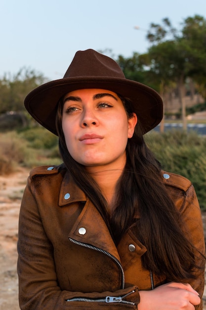 Giovane donna con cappello e giacca di pelle marrone che guarda in lontananza nel parco naturale