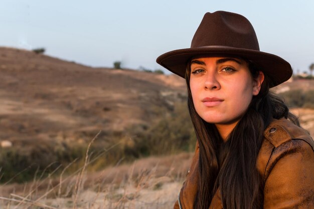 Giovane donna con cappello e giacca di pelle marrone che guarda la telecamera nel parco naturale