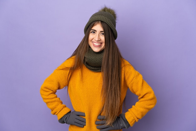 Giovane donna con cappello invernale isolato sulla parete viola in posa con le braccia all'anca e sorridente