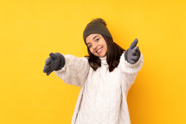 Giovane donna con cappello invernale sopra la parete gialla isolata che presenta e che invita a venire con la mano