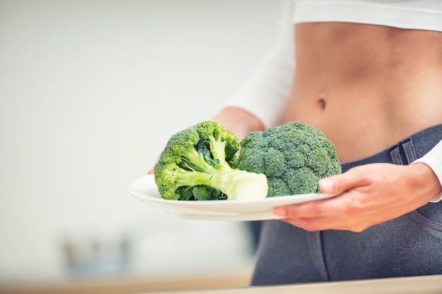 Giovane donna con figura sportiva in cucina che tiene tra le mani un piatto di broccoli.