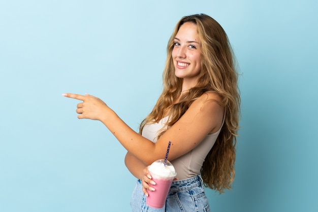 Giovane donna con frappè alla fragola isolato sulla parete blu che indica il dito a lato