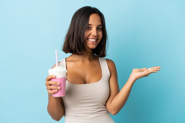 Giovane donna con frappè alla fragola isolato sulla parete blu che presenta un'idea mentre guarda sorridendo verso