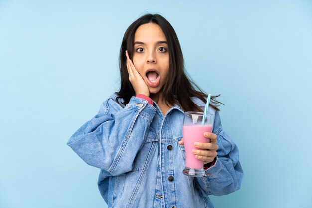 Giovane donna con frappè alla fragola sopra la parete blu con espressione facciale sorpresa e scioccata