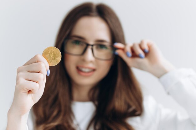 Giovane donna con gli occhiali in possesso di un Bitcoin isolato su sfondo grigio che fa un gesto di denaro