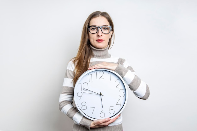 Giovane donna con gli occhiali in possesso di un orologio da parete su uno sfondo chiaro