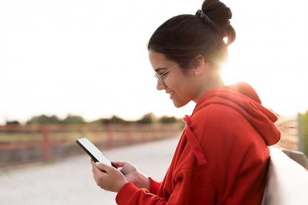 Giovane donna con gli occhiali sui suoi anni venti in chat sul suo telefono mentre era seduto in una panchina di un parco. Sta sorridendo e indossa un maglione rosso e una coda di cavallo. È retroilluminata con la luce del giorno al tramonto