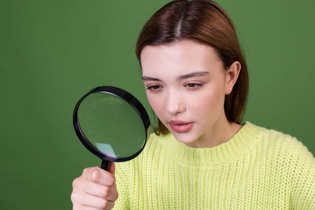 Giovane donna con grandi labbra marroni di trucco naturale perfetto in maglione casuale sulla parete verde con la ricerca della lente d'ingrandimento