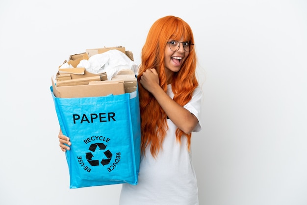 Giovane donna con i capelli arancioni che tiene in mano un sacchetto di riciclaggio pieno di carta da riciclare isolato su sfondo bianco che celebra una vittoria