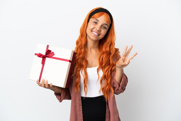 Giovane donna con i capelli arancioni che tiene un regalo isolato su sfondo bianco felice e conta tre con le dita