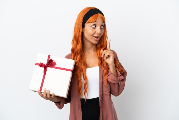 Giovane donna con i capelli arancioni che tiene un regalo isolato su sfondo bianco pensando a un'idea che punta il dito verso l'alto