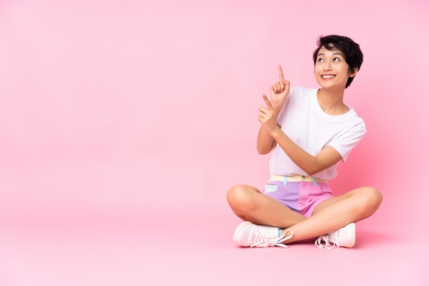 Giovane donna con i capelli corti, seduta sul pavimento sul muro rosa che puntava il dito indice una grande idea