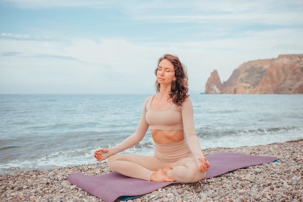 Giovane donna con i capelli neri, istruttore di fitness in leggings e top facendo stretching e pilates sulla stuoia di yoga vicino al mare. Concetto di routine di yoga fitness femminile Armonia e meditazione di uno stile di vita sano