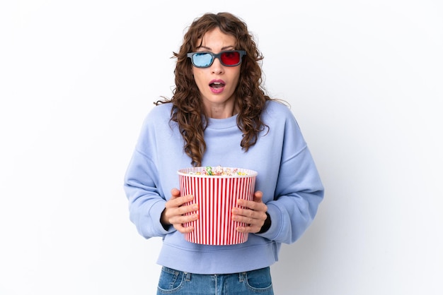 Giovane donna con i capelli ricci isolato su sfondo bianco sorpreso con occhiali 3d e con in mano un grande secchio di popcorn