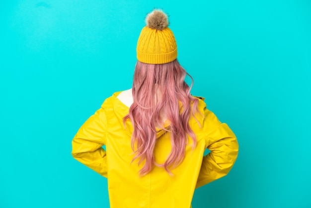 Giovane donna con i capelli rosa che indossa un cappotto antipioggia isolato su sfondo blu in posizione posteriore