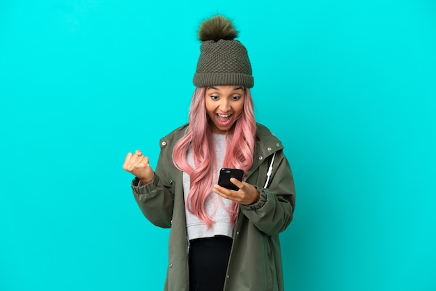 Giovane donna con i capelli rosa che indossa un cappotto antipioggia isolato su sfondo blu sorpreso e inviando un messaggio