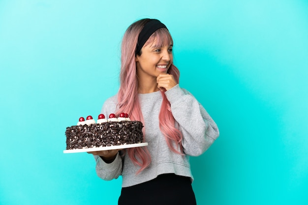 Giovane donna con i capelli rosa che tiene la torta di compleanno isolata su fondo blu che guarda al lato e che sorride