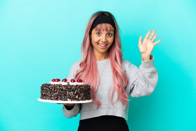 Giovane donna con i capelli rosa che tiene la torta di compleanno isolata su fondo blu che saluta con la mano con l'espressione felice