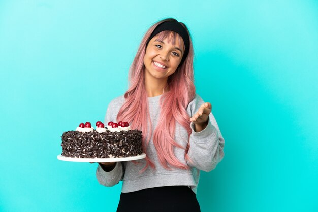 Giovane donna con i capelli rosa che tiene la torta di compleanno isolata su fondo blu che stringe la mano per aver chiuso un buon affare