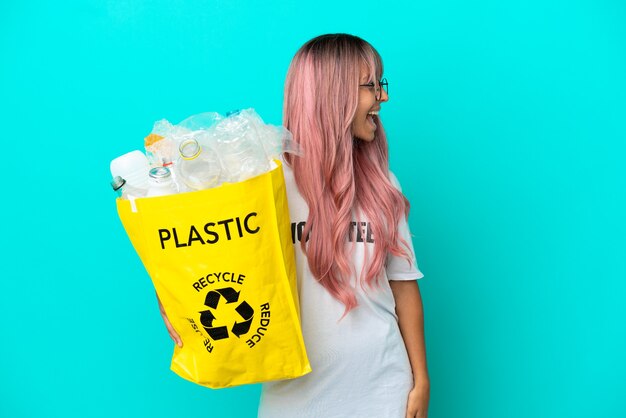 Giovane donna con i capelli rosa che tiene un sacchetto pieno di bottiglie di plastica da riciclare isolato su sfondo blu che ride in posizione laterale