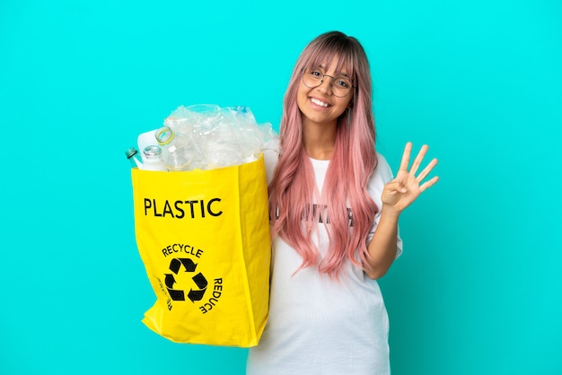 Giovane donna con i capelli rosa in possesso di un sacchetto pieno di bottiglie di plastica da riciclare isolato su sfondo blu felice e contando quattro con le dita