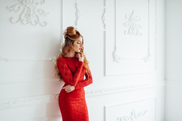 Giovane donna con i capelli rossi in un vestito rosso brillante in una stanza luminosa vicino a un muro bianco