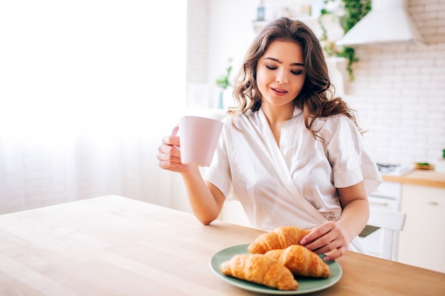 Giovane donna con i capelli scuri, seduto in cucina e bere caffè in mattinata. Prendendo un cornetto e sorridendo. Modello delizioso.