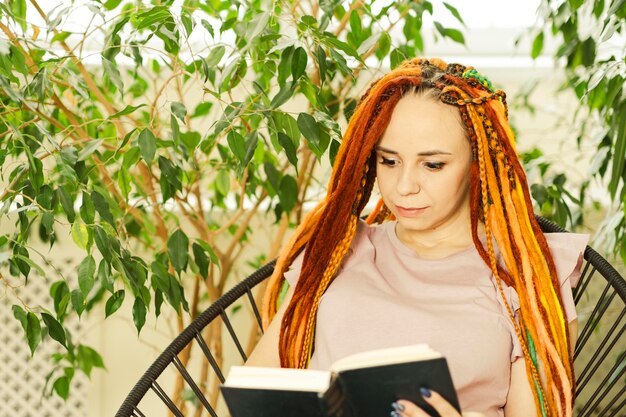 Giovane donna con i dreadlocks che legge il libro seduto su una sedia circondata da piante d'appartamento