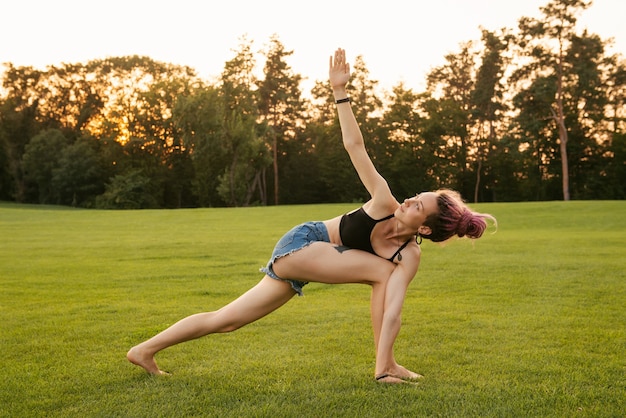 Giovane donna con i dreadlocks rosa che fa esercizi di yoga all'aperto e medita nel parco al tramonto