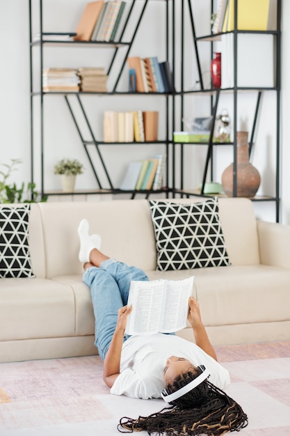 Giovane donna con i dreadlocks sdraiato sul pavimento con i piedi sul divano e leggere un libro interessante