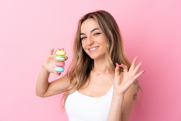 Giovane donna con i macarons sopra la parete rosa isolata che mostra segno giusto con le dita