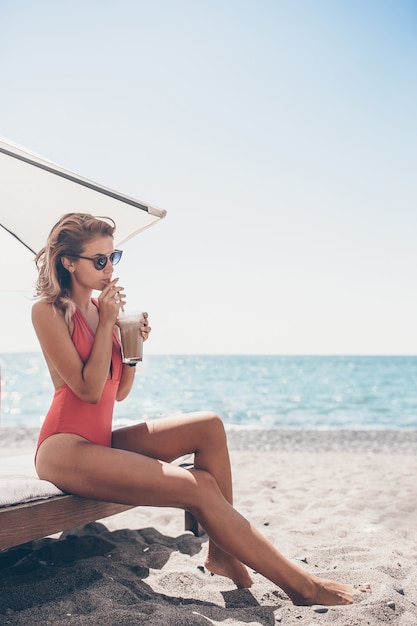 Giovane donna con il bicchiere da cocktail sulla spiaggia bianca che si siede sul lettino