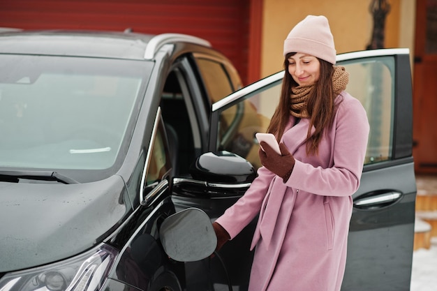 Giovane donna con il cellulare a portata di mano che carica la sua auto elettrica in inverno