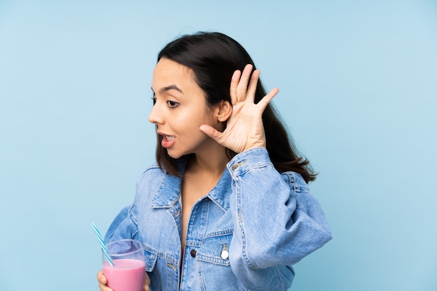 Giovane donna con il frappé della fragola sopra la parete blu isolata che ascolta qualcosa mettendo la mano sull'orecchio