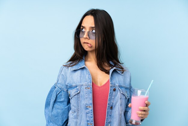 Giovane donna con il frappé della fragola sopra la parete blu isolata con l'espressione confusa del fronte