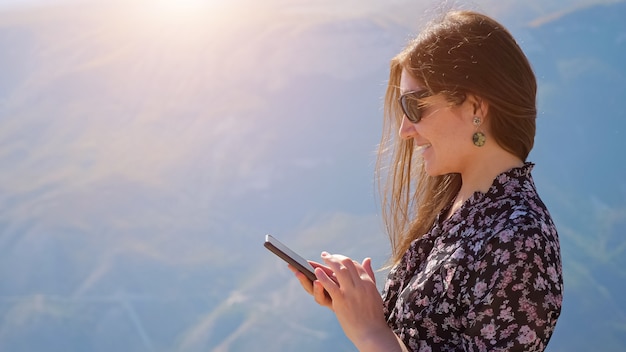 Giovane donna con il telefono sullo sfondo delle montagne in una giornata di sole, i capelli che fluttuano nel vento.