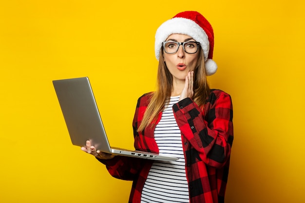 Giovane donna con la faccia sorpresa in cappello della Santa e computer portatile della holding della camicia a scacchi rossa