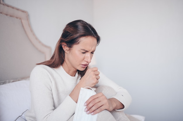 Giovane donna con la maschera malata dell'infezione virale dell'infezione virale di diffusione dell'infezione virale che si trova nell'isolamento di quarantena del letto a casa