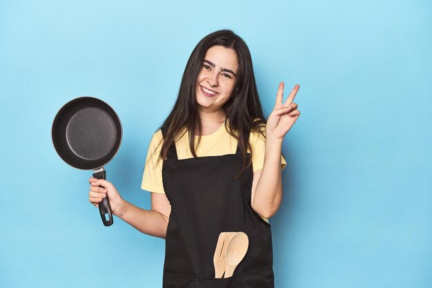 Giovane donna con la pentola sullo studio blu che mostra il numero due con le dita