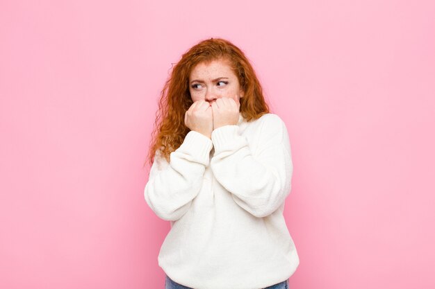 Giovane donna con la testa rossa che sembra preoccupata, ansiosa, stressata e impaurita, morde le unghie e cerca di copiare lo spazio laterale contro il muro rosa