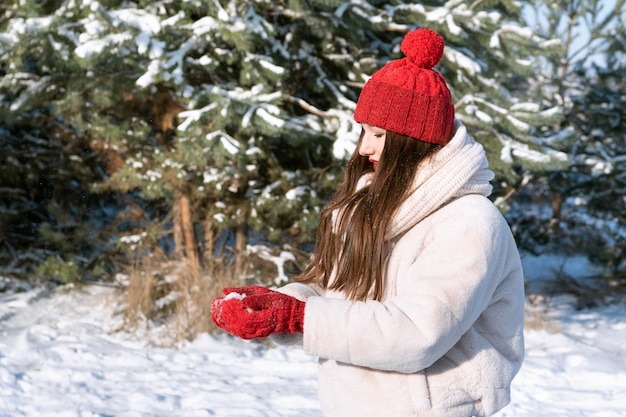 Giovane donna con labbra rosse in cappello rosso lavorato a maglia con la neve nelle mani nella foresta invernale Ragazza nel parco innevato