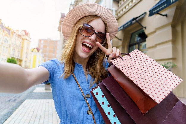 Giovane donna con le borse della spesa facendo selfie camminando in una città al giorno d'estate