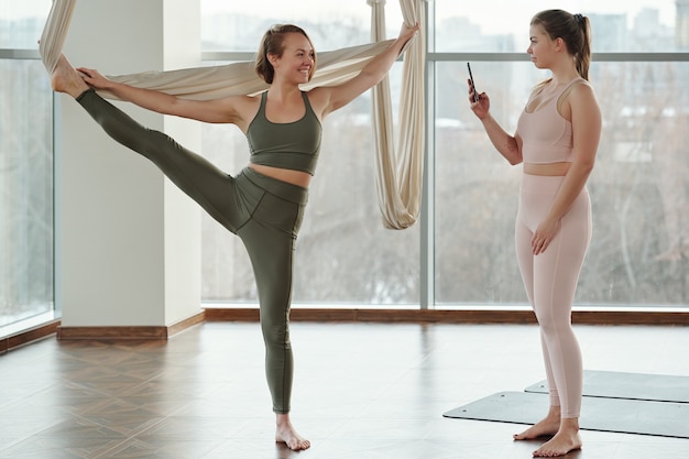 Giovane donna con lo smartphone che scatta foto di una donna bionda allegra in tuta da ginnastica che tiene un'amaca bianca durante l'esercizio di yoga a mosca