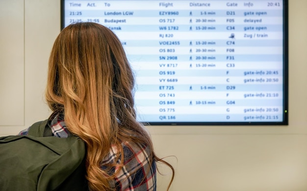 Giovane donna con lo zaino verde che guarda lo schermo del tabellone delle partenze all'aeroporto, vista da dietro.